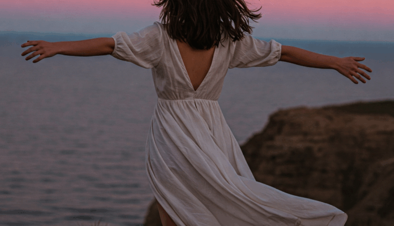 A woman stands on a cliff overlooking a calm ocean at sunset, symbolizing freedom and newfound peace after overcoming a toxic relationship. Her outstretched arms convey a sense of strength and liberation.
